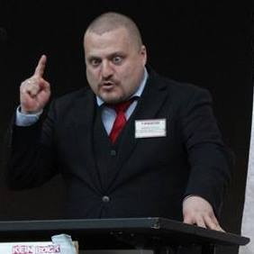 Im Anzug, mit erhobenem Zeigefinger und schielend eine Rede haltend.