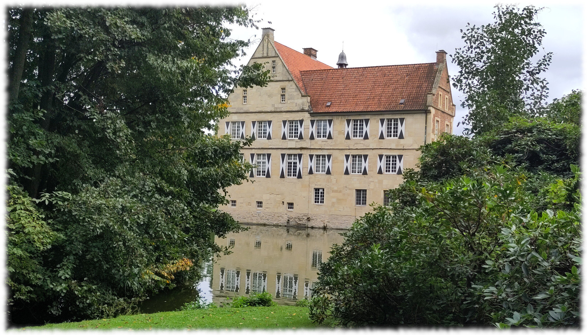 Burg Hlshoff mit Spiegelung im Wasser des Burggrabens, eingerahmt von Bumen und Struchern. Himmel bedeckt. 